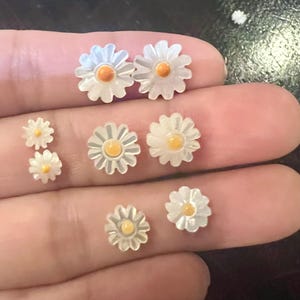 May include: A collection of daisy-shaped jewelry pieces, including earrings and small decorative elements. The daisy petals are white, and the centers are a bright orange-yellow. The pieces vary in size, with some appearing to be studs or small charms.
