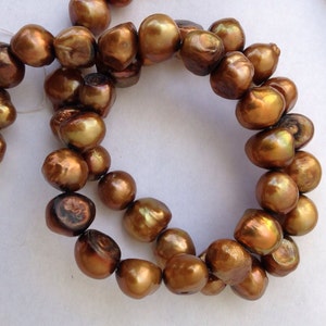 May include: A strand of brown baroque pearls with a golden sheen. The pearls are strung on a white thread and are arranged in a circular pattern.