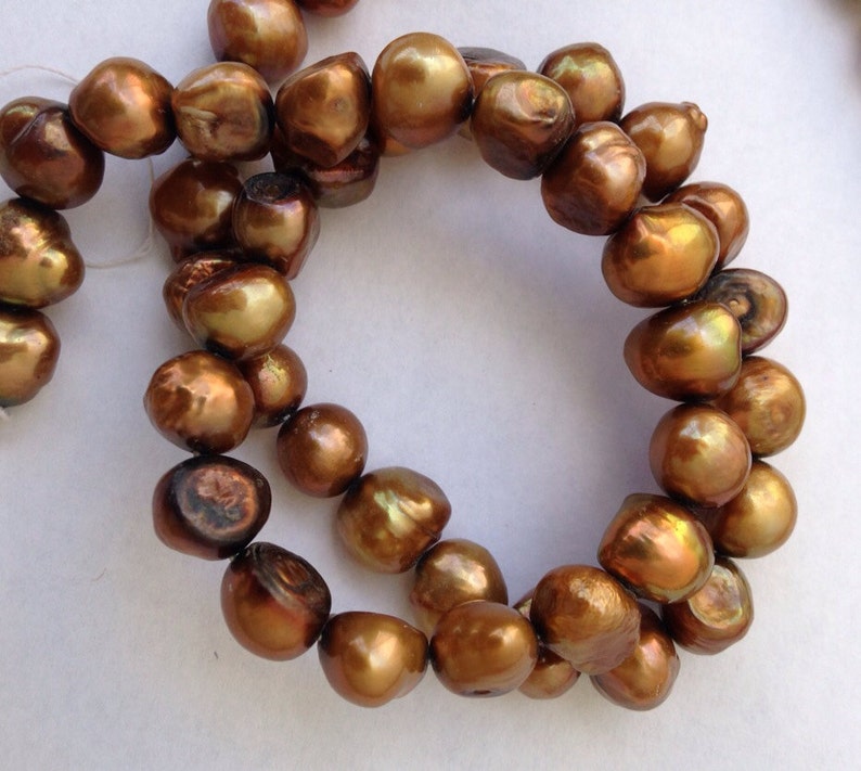 May include: A strand of brown baroque pearls with a golden sheen. The pearls are strung on a white thread and are arranged in a circular pattern.
