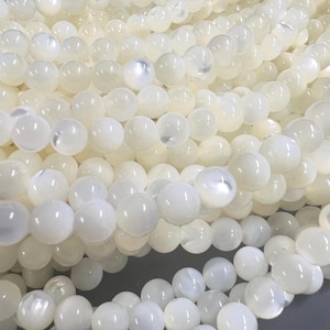 Puede incluir: Primer plano de varios hilos de cuentas redondas de color blanco roto. Las cuentas tienen un acabado nacarado, que refleja la luz. Las cuentas están ensartadas, creando una superficie fluida y texturizada. El efecto general es elegante y delicado.