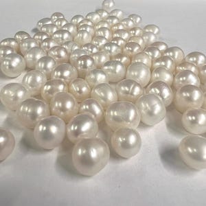 May include: A collection of lustrous, off-white pearls. The pearls are irregularly shaped, with a smooth, reflective surface. The pearls are scattered across a white surface, creating a sense of abundance and natural beauty. The pearls vary in size.