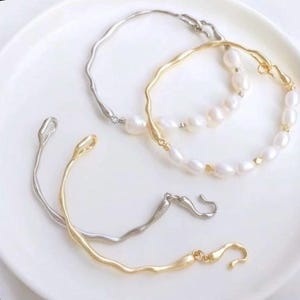 May include: Three delicate bracelets with a wavy design. Two of the bracelets are silver and gold, and the third bracelet is a pearl strand with silver and gold accents.