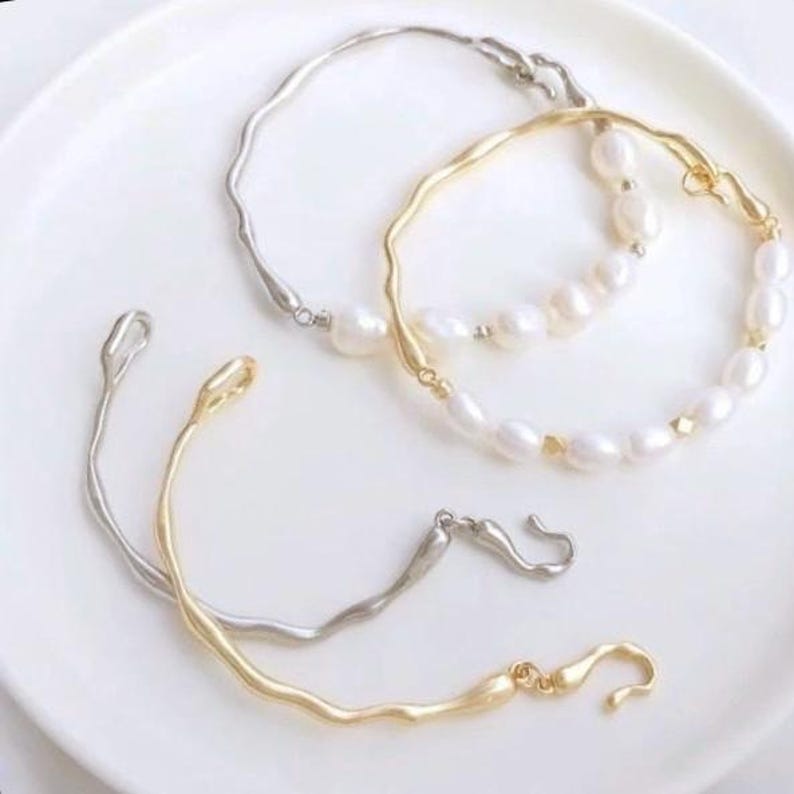May include: Three delicate bracelets with a wavy design. Two of the bracelets are silver and gold, and the third bracelet is a pearl strand with silver and gold accents.