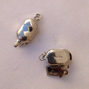May include: Two silver-toned metal clasps for a necklace or bracelet. One clasp has a small, decorative charm attached. The clasp on the right has the number "70271" engraved on it.