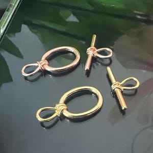 May include: Four metal toggle clasps in gold and rose gold tones. Two clasps are oval-shaped with a loop and knot design, and two are bar-shaped with a loop. The clasps are displayed on a reflective black surface.
