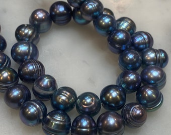 Puede incluir: Una hebra de perlas iridiscentes de color azul oscuro. Las perlas son redondas y tienen un acabado liso y brillante.