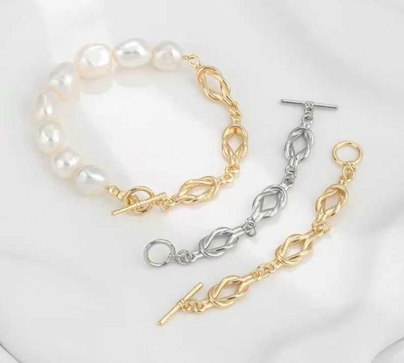 K&ouml;nnte beinhalten: Zwei Armb&auml;nder mit Knotendesign. Ein Armband ist golden und hat eine Reihe wei&szlig;er Perlen. Das andere Armband ist silber.