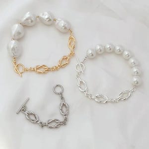 K&ouml;nnte beinhalten: Zwei Perlenarmb&auml;nder mit goldenen und silbernen Knotenverschl&uuml;ssen. Das goldene Armband hat gr&ouml;&szlig;ere Perlen und einen Schiebeverschluss. Das silberne Armband hat kleinere Perlen und einen T-Bar-Verschluss.