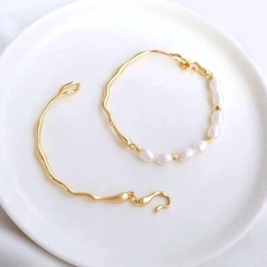 May include: Two gold bracelets, one with a simple wavy design and the other with a row of white pearls.