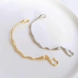 May include: Two simple, minimalist bracelets with a wavy design. One bracelet is gold and the other is silver.