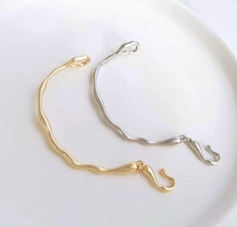 May include: Two simple, minimalist bracelets with a wavy design. One bracelet is gold and the other is silver.