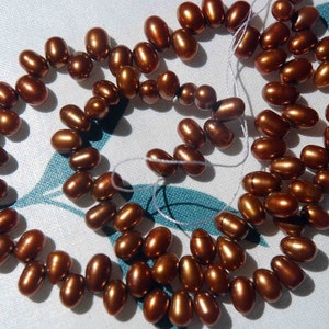 May include: A strand of brown pearl beads, strung on a white thread. The pearls are oval shaped and have a slightly iridescent sheen.