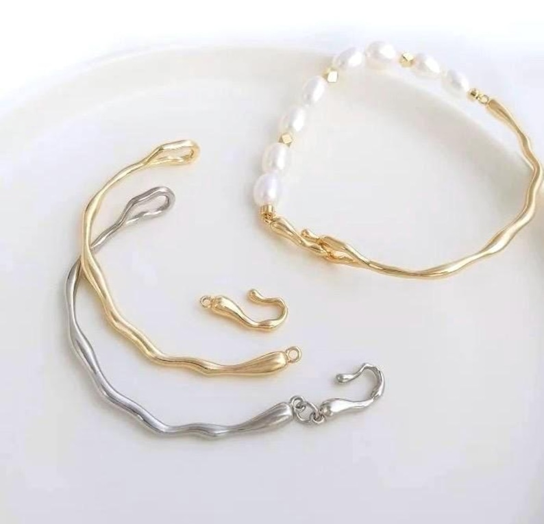 May include: Two gold and silver bracelets with a wavy design. The gold bracelet has a pearl and gold chain attached to it. There are two gold clasps.