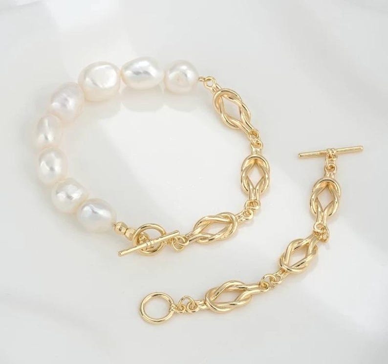 K&ouml;nnte beinhalten: Ein goldenes Kettenarmband mit Knotendesign und einer Reihe wei&szlig;er Perlen. Das Armband hat einen Schiebeverschluss.