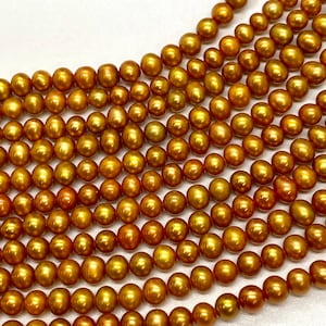 May include: A strand of golden pearls, with a warm, slightly reddish hue. The pearls are smooth and round, and they are arranged in a loose, flowing pattern.