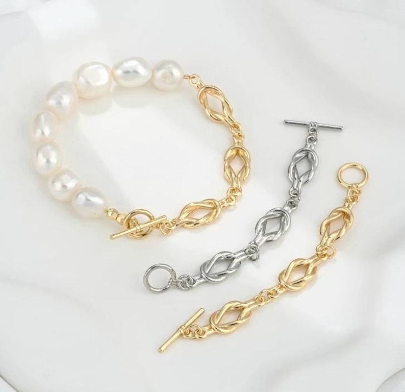 K&ouml;nnte beinhalten: Zwei Armb&auml;nder, eines mit einer goldenen Kette und einer Reihe wei&szlig;er Perlen, das andere mit einer silbernen Kette. Beide Armb&auml;nder haben einen Schiebeverschluss.