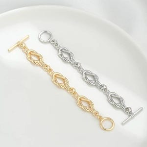 K&ouml;nnte beinhalten: Zwei Armb&auml;nder mit Knotendesign. Ein Armband ist golden und das andere silbern.