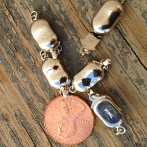 May include: A silver charm bracelet with a variety of silver charms, including a round charm with the inscription "925". The bracelet is lying on a wooden surface next to a US penny.
