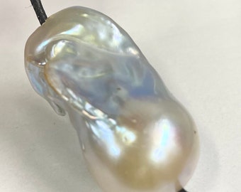 Pode incluir: Um pendente de pérola barroca iridescente grande em um cordão preto. A pérola tem uma superfície lisa e lustrosa com uma mistura de tons creme, brancos e azuis. A forma é irregular e orgânica, com formato de gota.