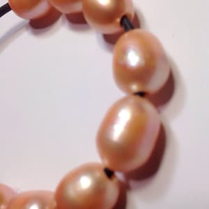 May include: A strand of peach-colored pearls strung on a black elastic cord. The pearls are oval-shaped and have a smooth, glossy finish.