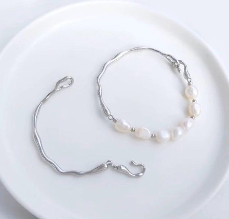 May include: Two silver bracelets with a delicate, irregular design. One bracelet features a row of white pearls.