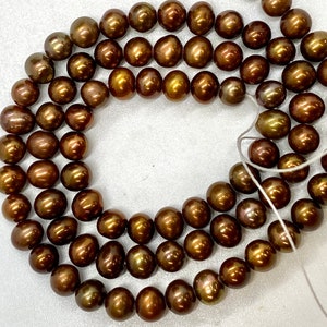 Könnte beinhalten: Eine Kette aus braunen Süßwasserperlen mit goldenem Schimmer. Die Perlen sind auf einem weißen Faden aufgefädelt und in einem Kreis angeordnet.