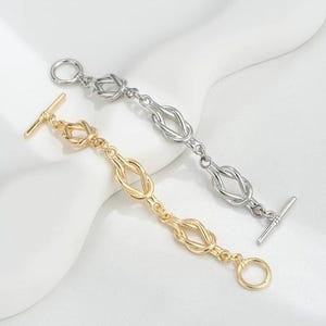 K&ouml;nnte beinhalten: Zwei Armb&auml;nder mit Knotendesign. Ein Armband ist golden und das andere silbern.