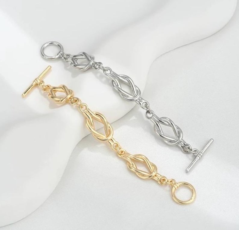 K&ouml;nnte beinhalten: Zwei Armb&auml;nder mit Knotendesign. Ein Armband ist golden und das andere silbern.