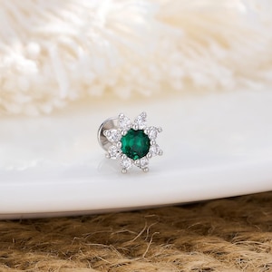Op de afbeelding: Een kleine, zilverkleurige oorbel met een centrale, ronde, smaragdgroene edelsteen. De edelsteen is omgeven door een halo van kleinere, heldere, ronde stenen, waardoor een sterrenpatroon ontstaat. De oorbel ligt op een wit oppervlak met een textuur achtergrond.