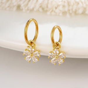 Puede incluir: Un par de pendientes de aro dorados con un dije en forma de flor. Cada dije presenta un grupo de piedras claras y brillantes. Los pendientes se exhiben sobre una superficie blanca, con un fondo suave y texturizado.