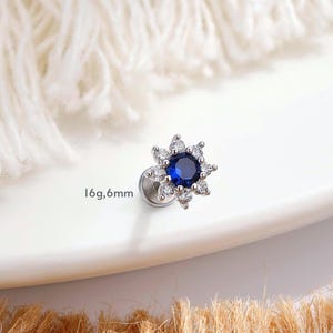 以下が含まれることがあります： 青い中心の石と周囲にクリアな石をあしらった、銀色のスタッドピアス。花のようなデザインです。「16g,6mm」の文字が見えます。直径は約6mmです。