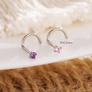 May include: Two nose rings with curved posts and square gemstones. One has a purple gemstone, the other a pink one. The text "20G 6mm" is visible. The jewellery is displayed on a white plate, with a neutral background.