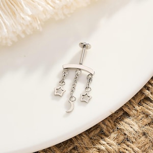 Peut inclure: Un piercing corporel argenté avec une barre incurvée et trois breloques pendantes : deux étoiles et un croissant de lune. Le bijou est présenté sur une assiette blanche avec un fond marron tissé.