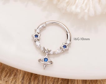 May include: A silver hoop earring with a dangling charm featuring a blue star and a small blue gem. The hoop is decorated with small white and blue gems and the text "16G 10mm" is visible.