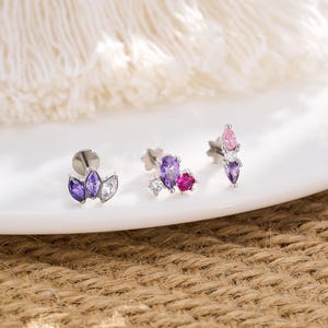 May include: Three pieces of jewellery with colourful gemstones. The pieces include a stud earring with purple and white stones, a stud earring with purple, pink, and white stones, and a stud earring with pink and purple stones. The jewellery is displayed on a white plate.