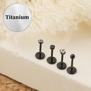 Puede incluir: Cinco piezas de joyería corporal de titanio negro con acentos de gemas transparentes. La joyería está dispuesta en una fila sobre una superficie blanca.