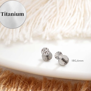 May include: Two silver titanium labret studs. One stud features a textured, geometric design, and the other has a radial pattern. The text "Titanium" is in a silver circle. The text "18G,6mm" is also visible.