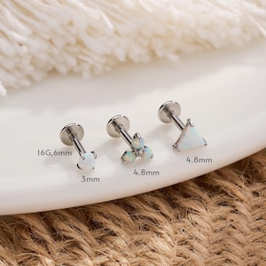 Puede incluir: Tres pendientes planos plateados con piedras blancas con forma de ópalo. Uno tiene forma de estrella, otro tiene tres piedras pequeñas y el tercero es un triángulo. Las medidas se indican en milímetros y calibre.