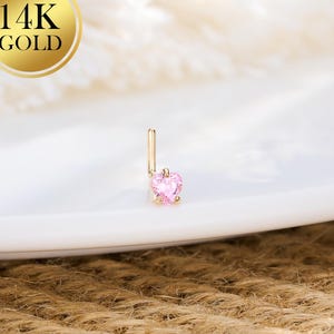 May include: A gold nose stud with a pink heart-shaped gemstone. The stud is set against a white background, with a gold 14K gold seal in the upper left corner. The jewelry is displayed on a white plate.