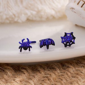 Puede incluir: Tres pendientes con temática de Halloween. Los pendientes son de color azul oscuro y presentan un diseño de araña, murciélago y telaraña. Los pendientes están sobre un plato blanco con una calavera en el fondo. El plato está sobre una superficie marrón.