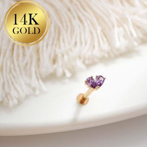 May include: A gold-colored, 14K gold lip piercing stud with two purple heart-shaped gemstones. The stud is on a white surface with a soft, white, textured background.