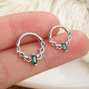 Puede incluir: Dos pendientes de aro plateados con un diseño similar a una vid y una piedra preciosa verde en el centro. Los pendientes son pequeños y delicados, con un acabado pulido. Las piedras preciosas son de forma ovalada y de un verde esmeralda intenso.