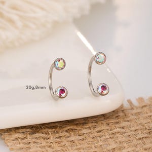 Könnte beinhalten: Ein Paar gebogene, silberfarbene Ohrringe. Jeder Ohrring hat oben einen klaren, irisierenden Edelstein und unten einen kleineren, roten Edelstein. Der Text "20g, 8mm" ist sichtbar. Die Ohrringe werden auf einer weißen Oberfläche präsentiert.