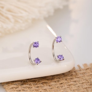 Peut inclure: Une paire de boucles d'oreilles argentées avec un design incurvé. Chaque boucle d'oreille comporte deux pierres précieuses carrées violettes. Les boucles d'oreilles sont présentées sur une surface blanche avec un fond neutre.