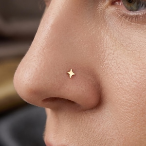 Pode incluir: Um pequeno piercing de nariz dourado em forma de estrela de quatro pontas. A joia está posicionada na lateral da narina e apresenta um acabamento metálico polido com pontas afiadas e simétricas que se estendem a partir de um centro.