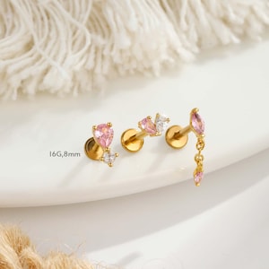 Puede incluir: Pendientes dorados con gemas rosas y blancas. Un pendiente presenta una gema rosa colgante. El texto "16G,8mm" es visible. Los pendientes se muestran sobre una superficie blanca con un fondo suave y texturizado.