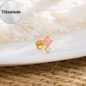May include: A gold-colored titanium labret stud with a pink gemstone and a small, clear crystal accent. The stud is designed for lip or ear piercings. The word "Titanium" is in a white circle in the background.