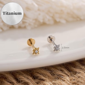 May include: Two titanium earrings with starburst designs. One is gold-toned, the other silver-toned. Each earring features a small, clear gem at the starburst's centre. The earrings are displayed on a white surface.