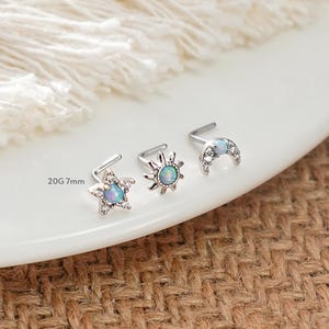 Puede incluir: Tres piercings de nariz plateados con diseños celestiales: una estrella, un sol y una luna creciente. Cada piercing presenta una piedra central similar a un ópalo y está adornado con piedras claras más pequeñas. El texto "20G 7mm" es visible.