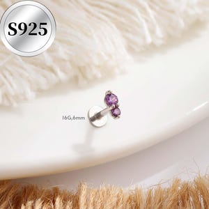 May include: A silver-colored lip piercing with two purple gemstones. The piercing is labeled "S925" and "16G,6mm". The jewelry is displayed on a white surface with a soft, textured background.